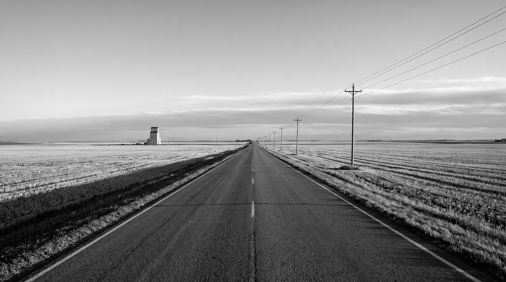 Generated black-and-white scene: a straight two-lane prairie highway between harvested fields, with power poles and a distant grain elevator; not a specific road.