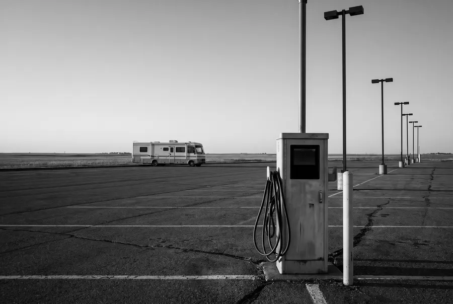 Generated black-and-white scene: a large parking lot at dusk with an electric vehicle charging pedestal in front and one motorhome parked far off; not a photograph of the casino lot.